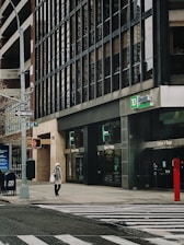 A city street scene featuring a TD Bank branch on the corner. A person is walking across a crosswalk, wearing a coat and a face mask. The building has large glass windows with the bank's logo visible. Nearby, there is a street sign indicating 'One Way' and a pedestrian crossing signal. A mailbox and other city street elements are also present.