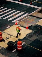 A person wearing a bright safety vest and helmet walks across a construction zone in an urban area. Orange traffic cones and barriers are placed around a section of the road, which appears to be under repair. The street is marked with white lines and other construction-related markings.