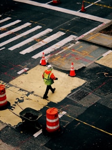 A person wearing a bright safety vest and helmet walks across a construction zone in an urban area. Orange traffic cones and barriers are placed around a section of the road, which appears to be under repair. The street is marked with white lines and other construction-related markings.
