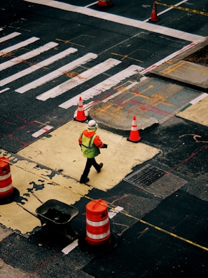 A person wearing a bright safety vest and helmet walks across a construction zone in an urban area. Orange traffic cones and barriers are placed around a section of the road, which appears to be under repair. The street is marked with white lines and other construction-related markings.