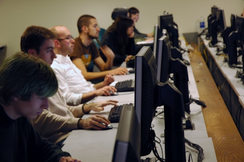 A group of people is seated in a row in front of computer monitors, focused on their tasks. The setting appears to be a computer lab or an office, with several monitors lined up on a long desk. The individuals are engrossed in their work, suggesting a study or work environment.