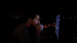 A person is intensely focused on a computer screen in a dimly lit environment, with light from the screen illuminating their face. Their hands are clasped near their face, suggesting deep concentration or contemplation.