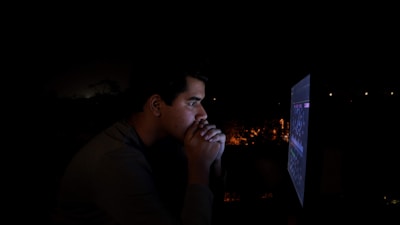A person is intensely focused on a computer screen in a dimly lit environment, with light from the screen illuminating their face. Their hands are clasped near their face, suggesting deep concentration or contemplation.