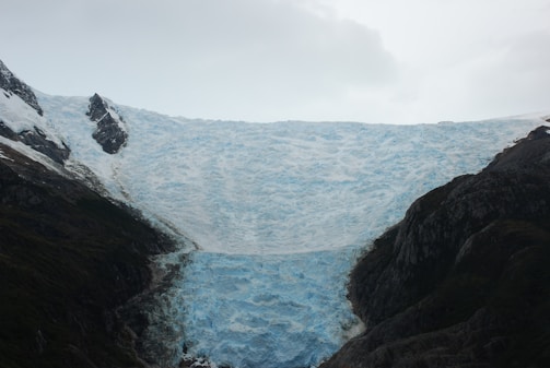 A massive glacier extends from between two rocky mountain peaks. The glacier is predominantly white with shades of blue, suggesting the compacted ice. Surrounding the glacier, the mountain terrain appears dark and rugged.