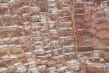 An aerial view of terraced salt ponds nestled in a mountainous landscape. The terraces are filled with mineral-rich saltwater, creating a patchwork pattern of earthy and whitish hues. The terrain appears rugged, with a mix of smooth and jagged textures.