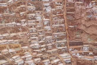 An aerial view of terraced salt ponds nestled in a mountainous landscape. The terraces are filled with mineral-rich saltwater, creating a patchwork pattern of earthy and whitish hues. The terrain appears rugged, with a mix of smooth and jagged textures.