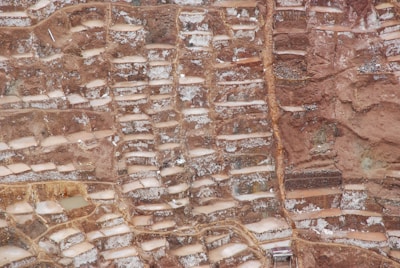 An aerial view of terraced salt ponds nestled in a mountainous landscape. The terraces are filled with mineral-rich saltwater, creating a patchwork pattern of earthy and whitish hues. The terrain appears rugged, with a mix of smooth and jagged textures.