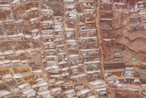 An aerial view of terraced salt ponds nestled in a mountainous landscape. The terraces are filled with mineral-rich saltwater, creating a patchwork pattern of earthy and whitish hues. The terrain appears rugged, with a mix of smooth and jagged textures.