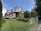 A large, traditional wooden house sits on a small hill surrounded by lush greenery and landscaped gardens. The house features steeply pitched roofs with multiple brick chimneys and green-striped awnings. A stone path leads up to the entrance, bordered by neatly trimmed bushes and a stone wall.