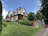 A large, traditional wooden house sits on a small hill surrounded by lush greenery and landscaped gardens. The house features steeply pitched roofs with multiple brick chimneys and green-striped awnings. A stone path leads up to the entrance, bordered by neatly trimmed bushes and a stone wall.