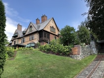 A large, traditional wooden house sits on a small hill surrounded by lush greenery and landscaped gardens. The house features steeply pitched roofs with multiple brick chimneys and green-striped awnings. A stone path leads up to the entrance, bordered by neatly trimmed bushes and a stone wall.