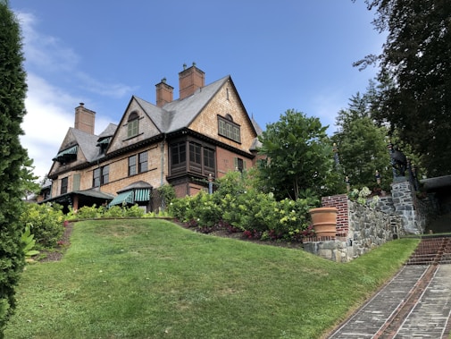 A large, traditional wooden house sits on a small hill surrounded by lush greenery and landscaped gardens. The house features steeply pitched roofs with multiple brick chimneys and green-striped awnings. A stone path leads up to the entrance, bordered by neatly trimmed bushes and a stone wall.