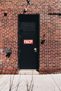 A black door with a red and white 'FACP' sign is set into a brick wall. The wall features a combination of brown and red bricks with contrasting white mortar. Above the door are two security cameras, and near the door handle, there is a small caution sign.
