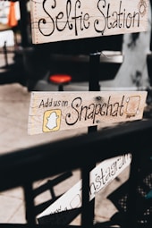 Wooden signs with handwritten text advertise social media engagement, including platforms like Snapchat and Instagram, attached to a pole in a casual outdoor setting. The text is decorative, and the imagery provides a vintage, handcrafted feel.