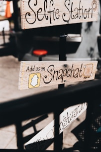 Wooden signs with handwritten text advertise social media engagement, including platforms like Snapchat and Instagram, attached to a pole in a casual outdoor setting. The text is decorative, and the imagery provides a vintage, handcrafted feel.