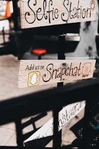 Wooden signs with handwritten text advertise social media engagement, including platforms like Snapchat and Instagram, attached to a pole in a casual outdoor setting. The text is decorative, and the imagery provides a vintage, handcrafted feel.