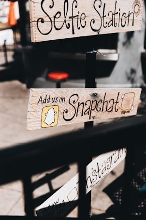 Wooden signs with handwritten text advertise social media engagement, including platforms like Snapchat and Instagram, attached to a pole in a casual outdoor setting. The text is decorative, and the imagery provides a vintage, handcrafted feel.