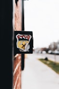 A close-up view of a black mailbox affixed to a red brick wall. The mailbox features a colorful emblem featuring mountains, a yellow road, and a pickaxe. The background is slightly blurred, showing a sidewalk and street with some greenery and indistinct buildings.