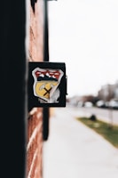 A close-up view of a black mailbox affixed to a red brick wall. The mailbox features a colorful emblem featuring mountains, a yellow road, and a pickaxe. The background is slightly blurred, showing a sidewalk and street with some greenery and indistinct buildings.