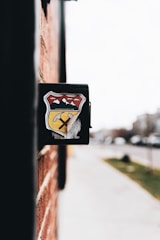 A close-up view of a black mailbox affixed to a red brick wall. The mailbox features a colorful emblem featuring mountains, a yellow road, and a pickaxe. The background is slightly blurred, showing a sidewalk and street with some greenery and indistinct buildings.