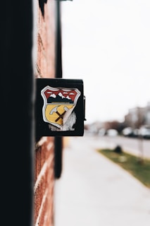 A close-up view of a black mailbox affixed to a red brick wall. The mailbox features a colorful emblem featuring mountains, a yellow road, and a pickaxe. The background is slightly blurred, showing a sidewalk and street with some greenery and indistinct buildings.