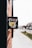 A close-up view of a black mailbox affixed to a red brick wall. The mailbox features a colorful emblem featuring mountains, a yellow road, and a pickaxe. The background is slightly blurred, showing a sidewalk and street with some greenery and indistinct buildings.