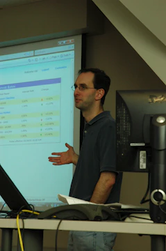 A person standing in front of a projector screen displaying interest rates and other financial information. The person appears to be giving a presentation or lecture in a classroom or office setting. A computer monitor and some papers are visible on a desk in the foreground.
