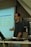 A person standing in front of a projector screen displaying interest rates and other financial information. The person appears to be giving a presentation or lecture in a classroom or office setting. A computer monitor and some papers are visible on a desk in the foreground.