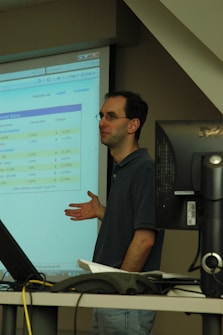 A person standing in front of a projector screen displaying interest rates and other financial information. The person appears to be giving a presentation or lecture in a classroom or office setting. A computer monitor and some papers are visible on a desk in the foreground.