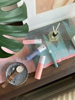 A collection of personal care items and accessories arranged on a table. There are several colorful bottles, including pink, blue, and teal perfumes, positioned on a multicolored woven mat. A glass diffuser with reeds is also visible. To the side, a small striped dish holds a gold watch and a decorative item. A large green leaf partially frames the scene.