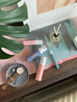 A collection of personal care items and accessories arranged on a table. There are several colorful bottles, including pink, blue, and teal perfumes, positioned on a multicolored woven mat. A glass diffuser with reeds is also visible. To the side, a small striped dish holds a gold watch and a decorative item. A large green leaf partially frames the scene.