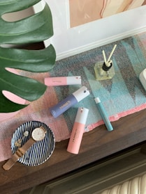 A collection of personal care items and accessories arranged on a table. There are several colorful bottles, including pink, blue, and teal perfumes, positioned on a multicolored woven mat. A glass diffuser with reeds is also visible. To the side, a small striped dish holds a gold watch and a decorative item. A large green leaf partially frames the scene.