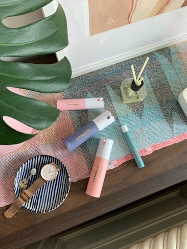 A collection of personal care items and accessories arranged on a table. There are several colorful bottles, including pink, blue, and teal perfumes, positioned on a multicolored woven mat. A glass diffuser with reeds is also visible. To the side, a small striped dish holds a gold watch and a decorative item. A large green leaf partially frames the scene.