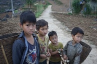 Four young boys with serious expressions stand on a narrow dirt path in a rural setting. Each boy carries a woven basket on their back, likely used for gathering or transporting goods. They wear casual, somewhat worn clothing. The background features a cultivated field with earthy tones and scattered greenery, accented by a few simple structures and a mountainous landscape.