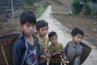 Four young boys with serious expressions stand on a narrow dirt path in a rural setting. Each boy carries a woven basket on their back, likely used for gathering or transporting goods. They wear casual, somewhat worn clothing. The background features a cultivated field with earthy tones and scattered greenery, accented by a few simple structures and a mountainous landscape.