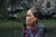 An older woman is outdoors, seated near a rocky and natural landscape. She appears to be covering part of her face with her hand, possibly in thought or emotion. Her hair is tied back, and she is wearing a jacket over a pink shirt.
