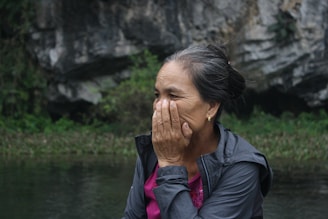 An older woman is outdoors, seated near a rocky and natural landscape. She appears to be covering part of her face with her hand, possibly in thought or emotion. Her hair is tied back, and she is wearing a jacket over a pink shirt.
