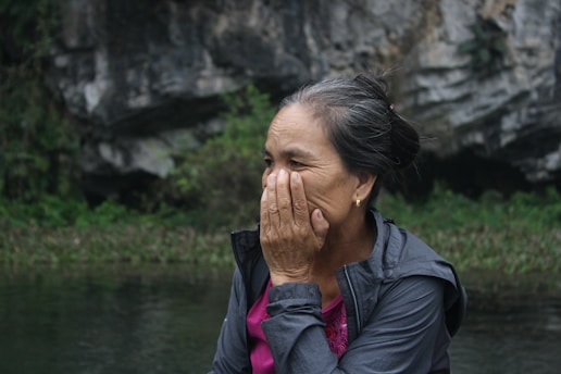 An older woman is outdoors, seated near a rocky and natural landscape. She appears to be covering part of her face with her hand, possibly in thought or emotion. Her hair is tied back, and she is wearing a jacket over a pink shirt.