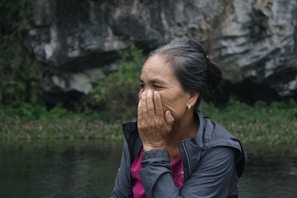 An older woman is outdoors, seated near a rocky and natural landscape. She appears to be covering part of her face with her hand, possibly in thought or emotion. Her hair is tied back, and she is wearing a jacket over a pink shirt.