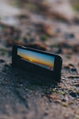 A mobile phone positioned on sandy ground capturing a scenic view of a sunset over the horizon, displaying a vibrant orange and blue sky on its screen. The phone is slightly angled, highlighting the rugged texture of the sand around it.