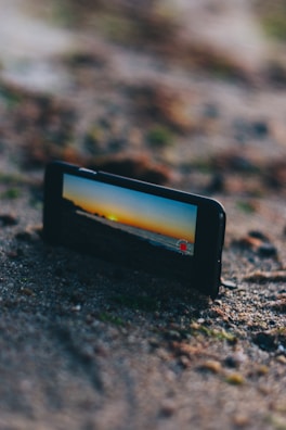 A mobile phone positioned on sandy ground capturing a scenic view of a sunset over the horizon, displaying a vibrant orange and blue sky on its screen. The phone is slightly angled, highlighting the rugged texture of the sand around it.
