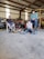 A workshop setting features two men seated on chairs, one wearing a hat and the other a cap. A young person lies on the ground with a dog next to them. Tools, a red vehicle, and various workshop items are in the background.