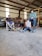 A workshop setting features two men seated on chairs, one wearing a hat and the other a cap. A young person lies on the ground with a dog next to them. Tools, a red vehicle, and various workshop items are in the background.