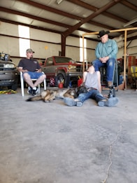 A workshop setting features two men seated on chairs, one wearing a hat and the other a cap. A young person lies on the ground with a dog next to them. Tools, a red vehicle, and various workshop items are in the background.
