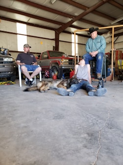 A workshop setting features two men seated on chairs, one wearing a hat and the other a cap. A young person lies on the ground with a dog next to them. Tools, a red vehicle, and various workshop items are in the background.