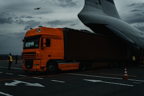 An orange truck is positioned on an airport runway, with its trailer partially inside a large cargo plane. The sky is overcast, conveying a moody atmosphere. A person wearing a high-visibility vest stands in the foreground, and another person is near the airplane. An airplane is flying above in the distant sky.