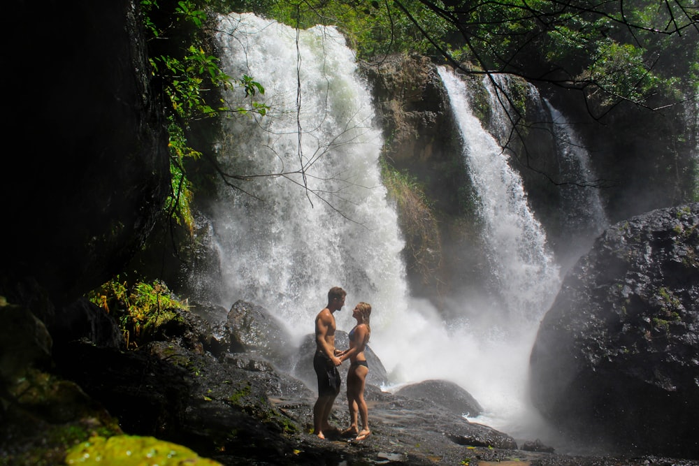 2 women in black tank top and black shorts standing on rock near waterfalls during daytime