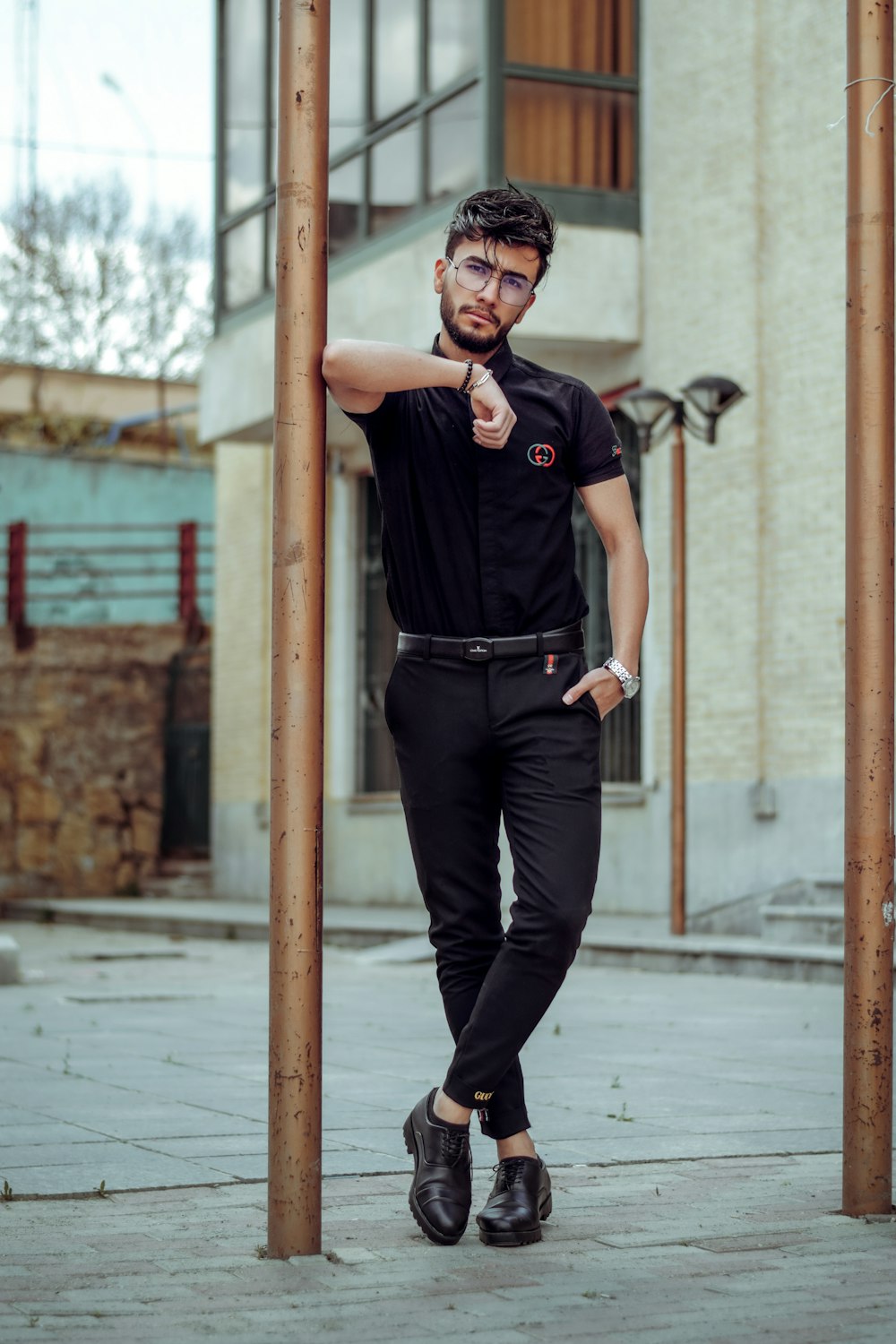 uomo in polo nera e pantaloni neri che indossa occhiali da sole neri in piedi sul marciapiede durante il giorno