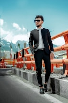 A man in a stylish black suit and sunglasses stands confidently beside an orange safety barrier on a road. The background features a mountainous landscape under a clear blue sky.