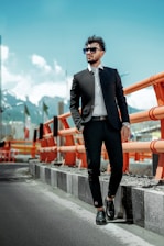 A man in a stylish black suit and sunglasses stands confidently beside an orange safety barrier on a road. The background features a mountainous landscape under a clear blue sky.